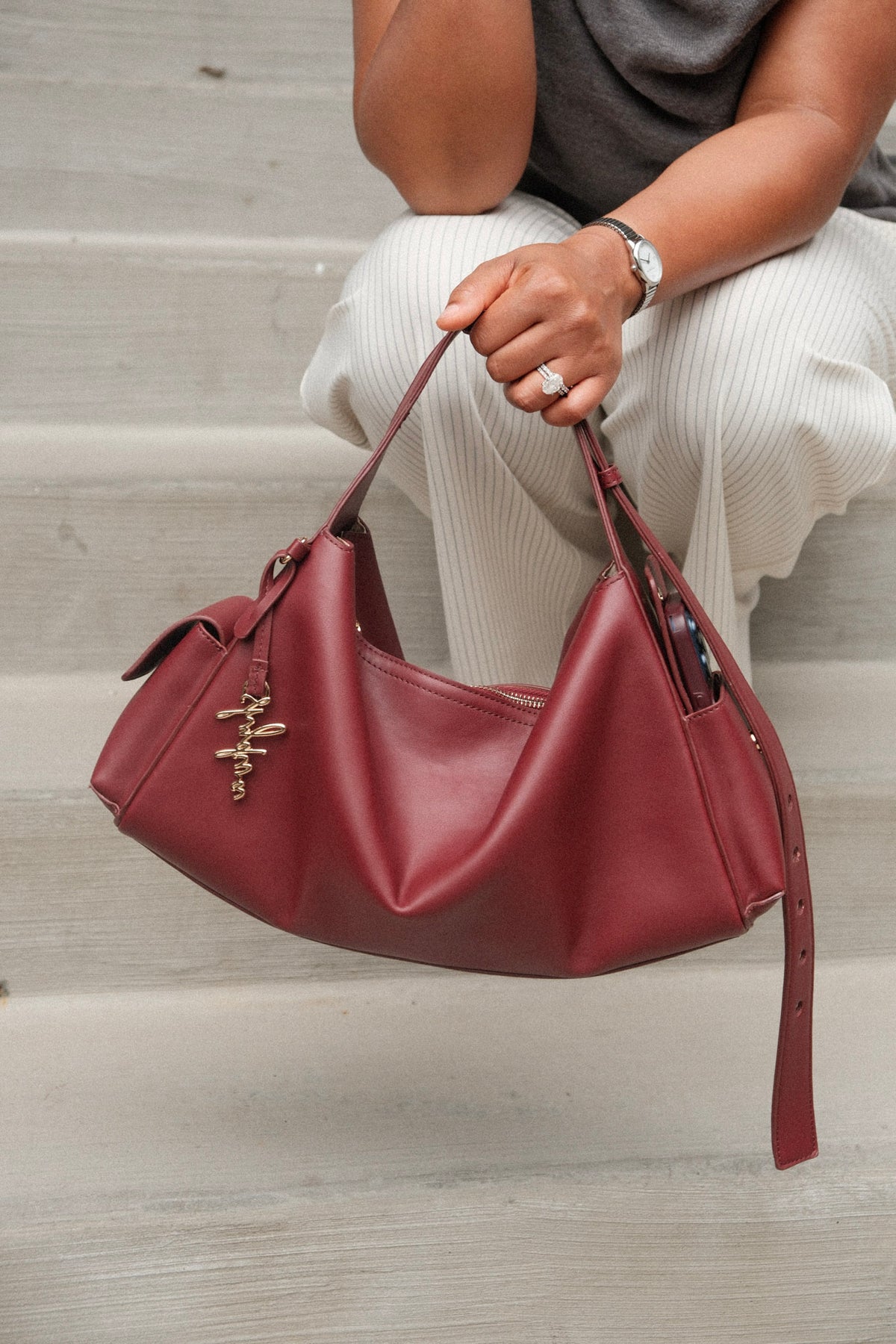 A burgundy faux-leather slouchy shoulder bag with gold-tone hardware and a hanging script charm, held by a person sitting on concrete steps.