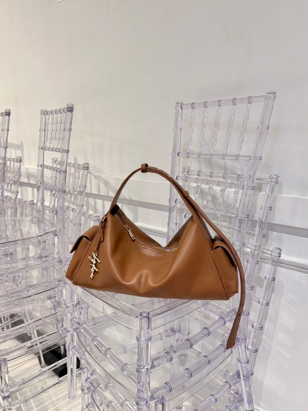 MILA cognac shoulder bag with gold-tone logo charm resting on a clear chair.