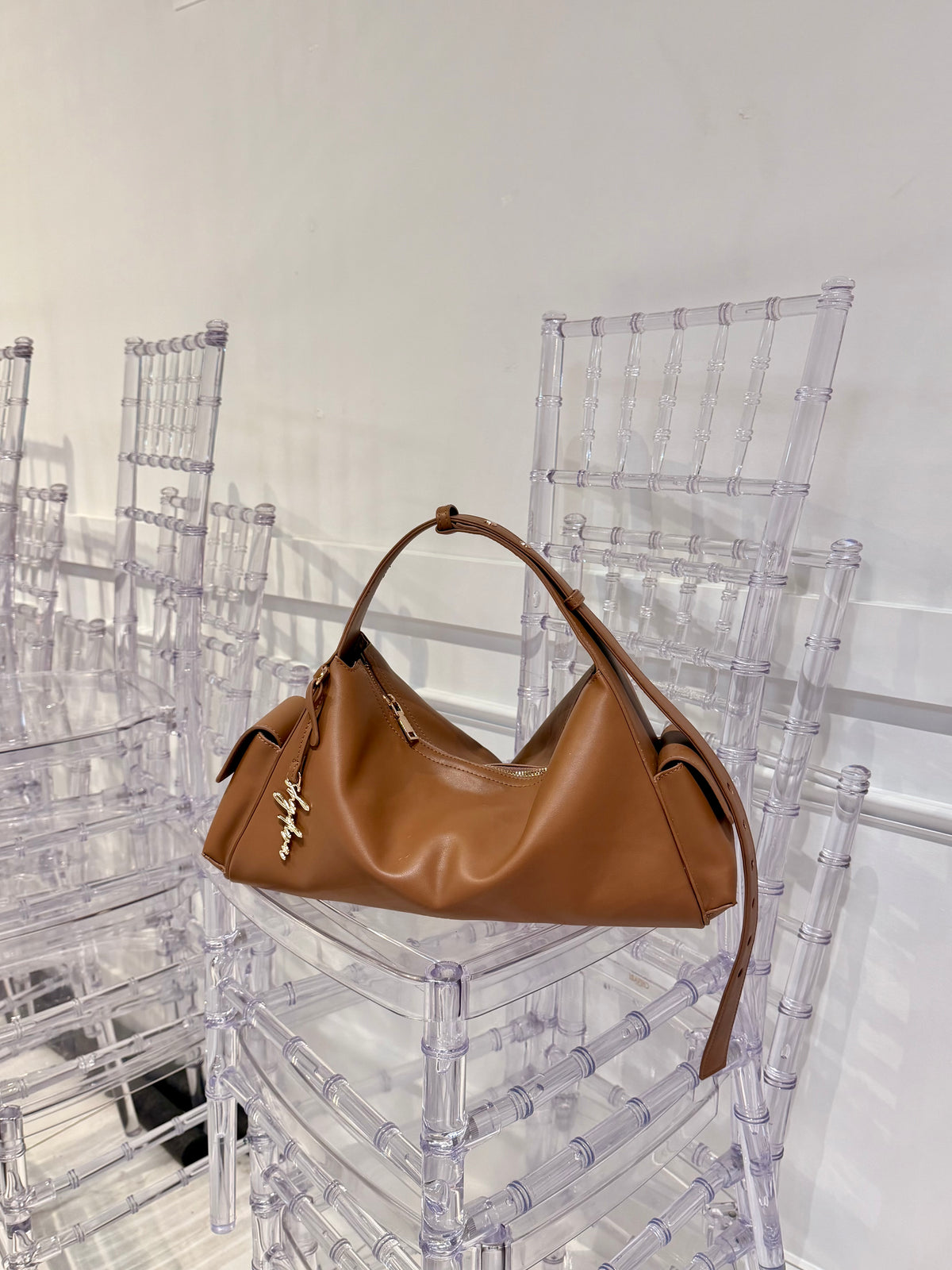 MILA cognac shoulder bag with gold-tone logo charm resting on a clear chair.