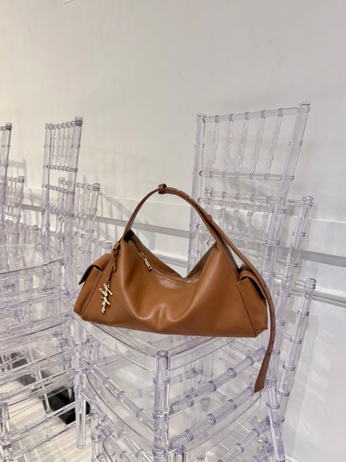 MILA cognac shoulder bag with gold-tone logo charm resting on a clear chair.